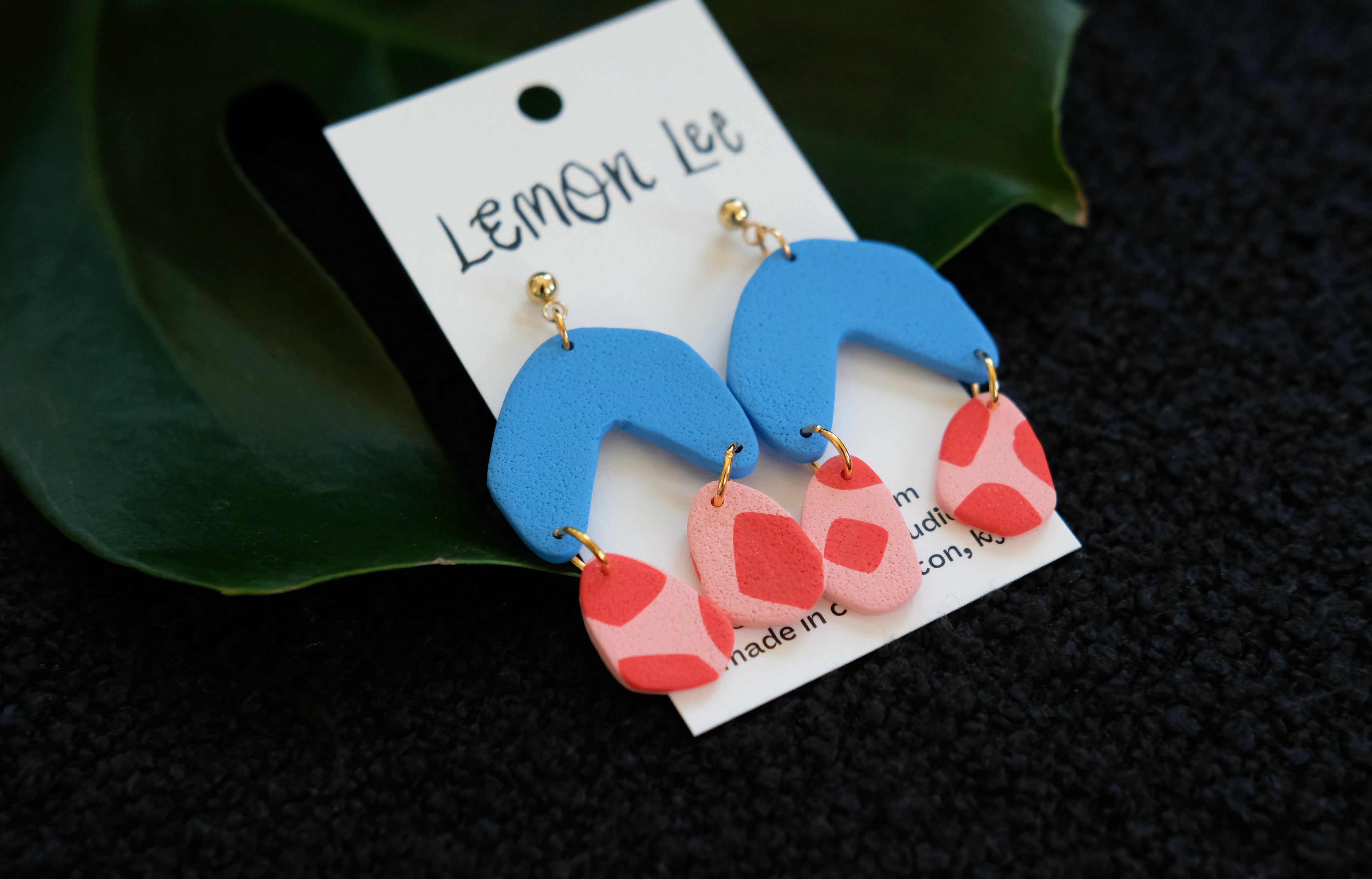 Blue and pink earrings on a branded card against a dark background