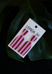 Pink and brown striped earrings on a branded card against a dark leafy background