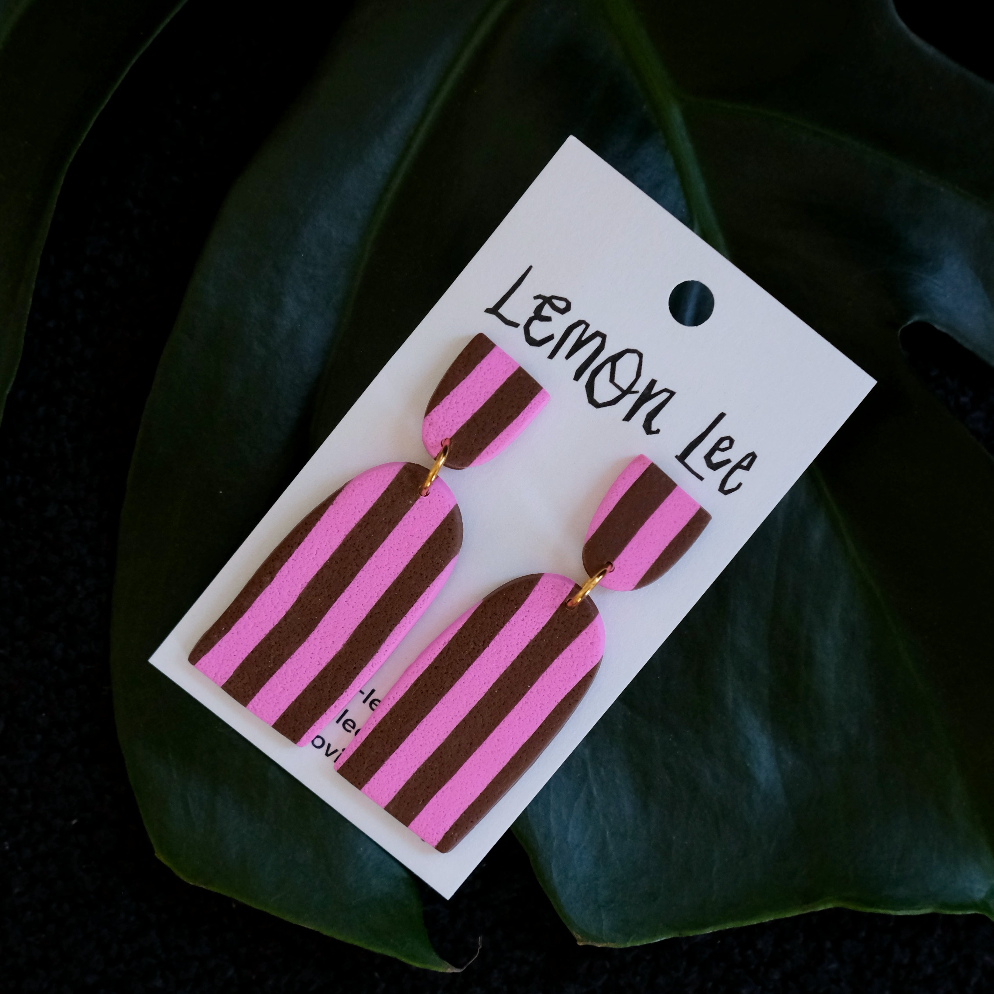 Pink and brown striped earrings on a branded card against a dark leafy background