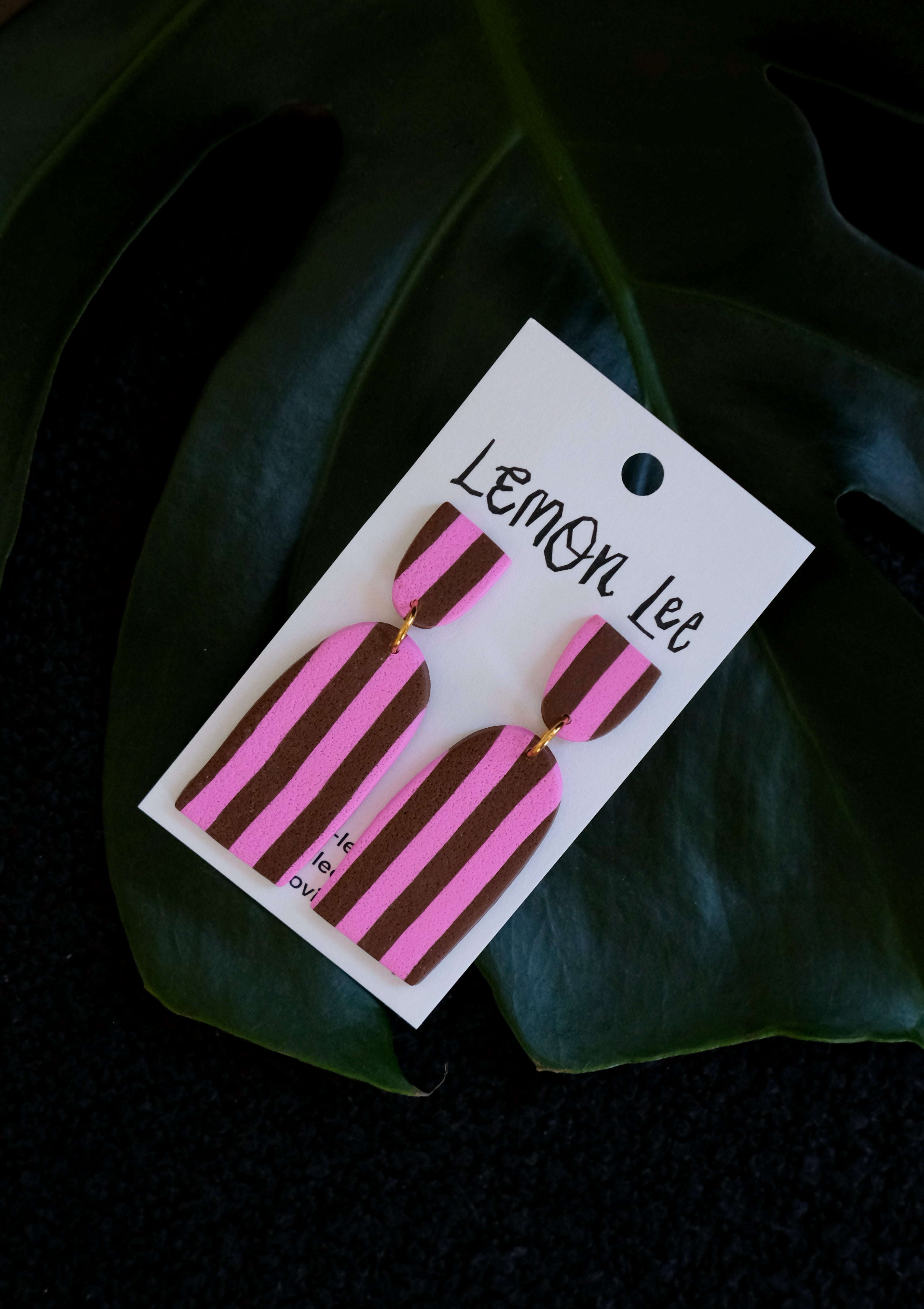 Pink and brown striped earrings on a branded card against a dark leafy background
