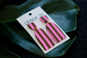 Pair of pink and brown striped earrings on a branded card, set against a dark background with green leaves.