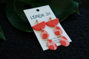 Red and pink earrings on a branded card with green leaves in the background