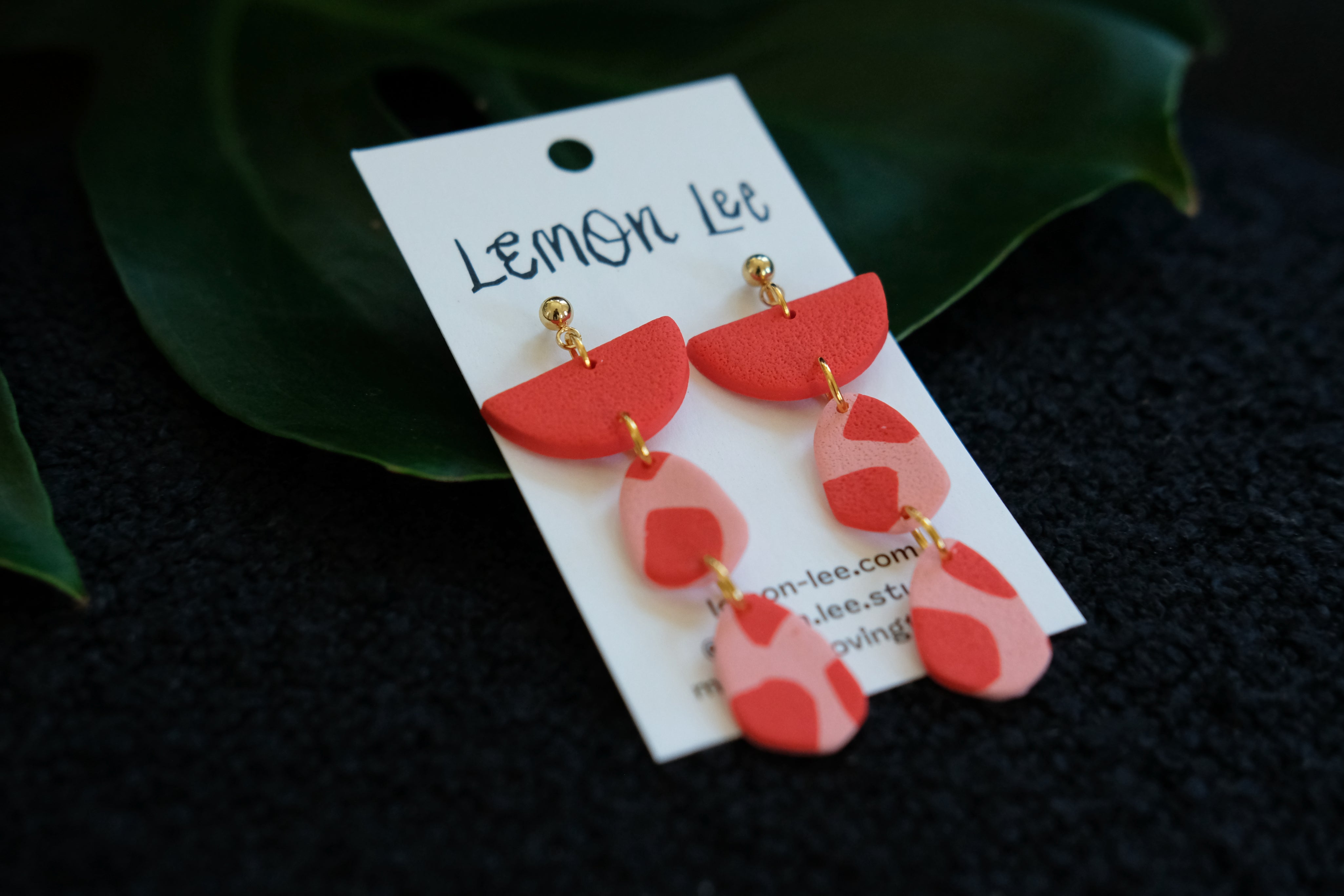 Red and pink earrings on a branded card with green leaves in the background